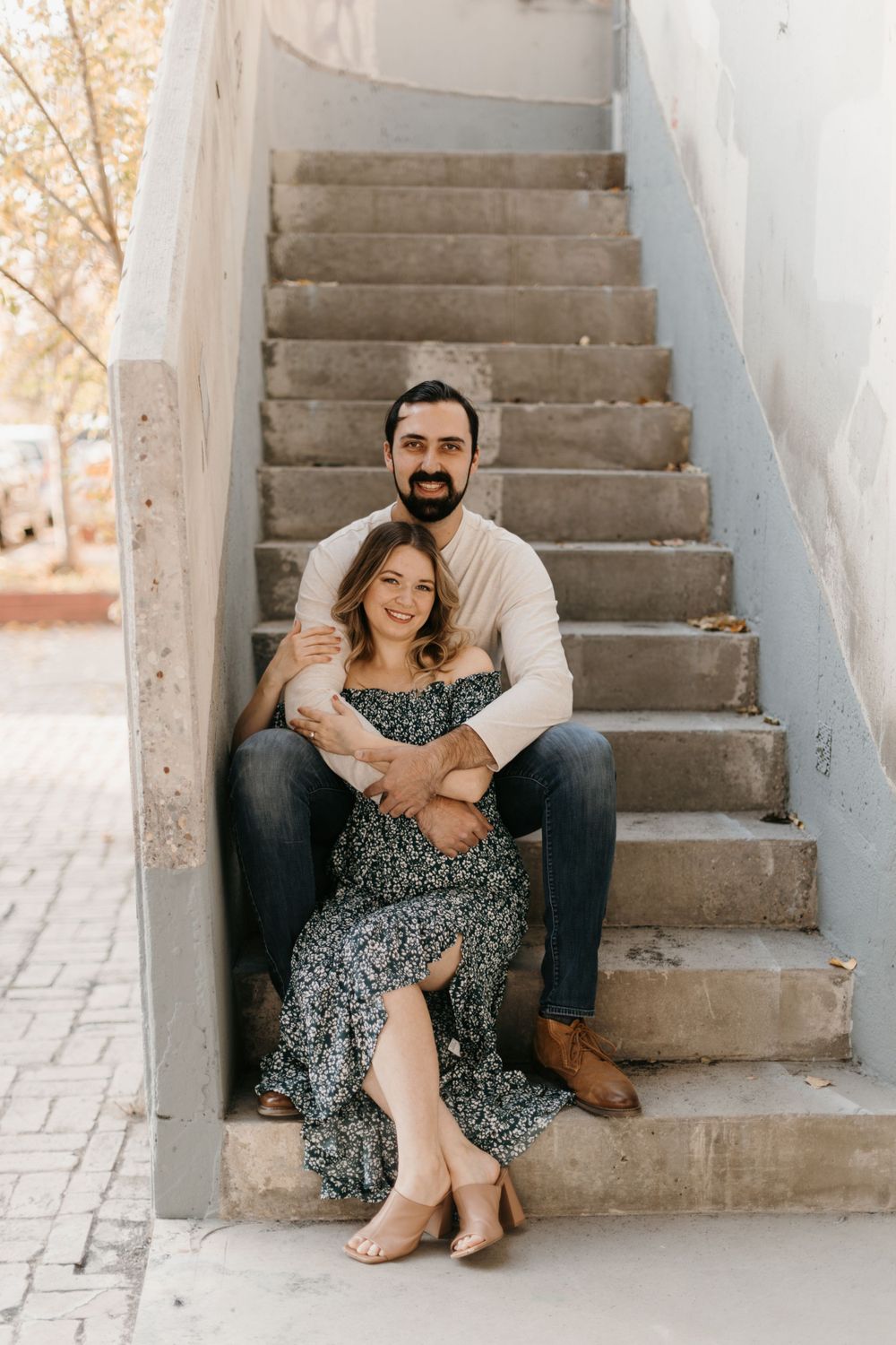 A couple sits on concrete stairs, man embracing woman in floral dress, both smiling at camera.