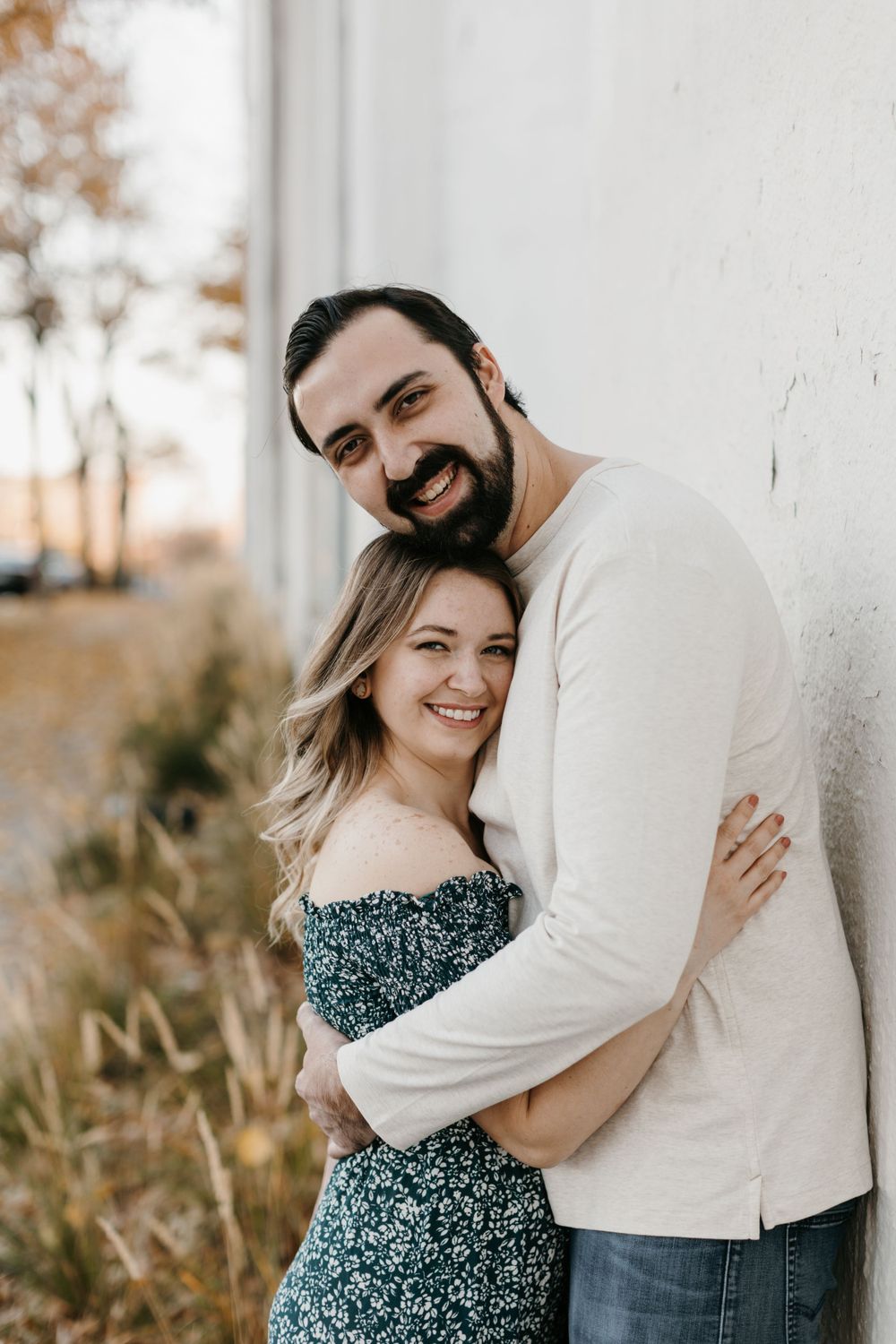 Smiling bearded man embracing woman in green floral dress against white wall in golden autumn light.