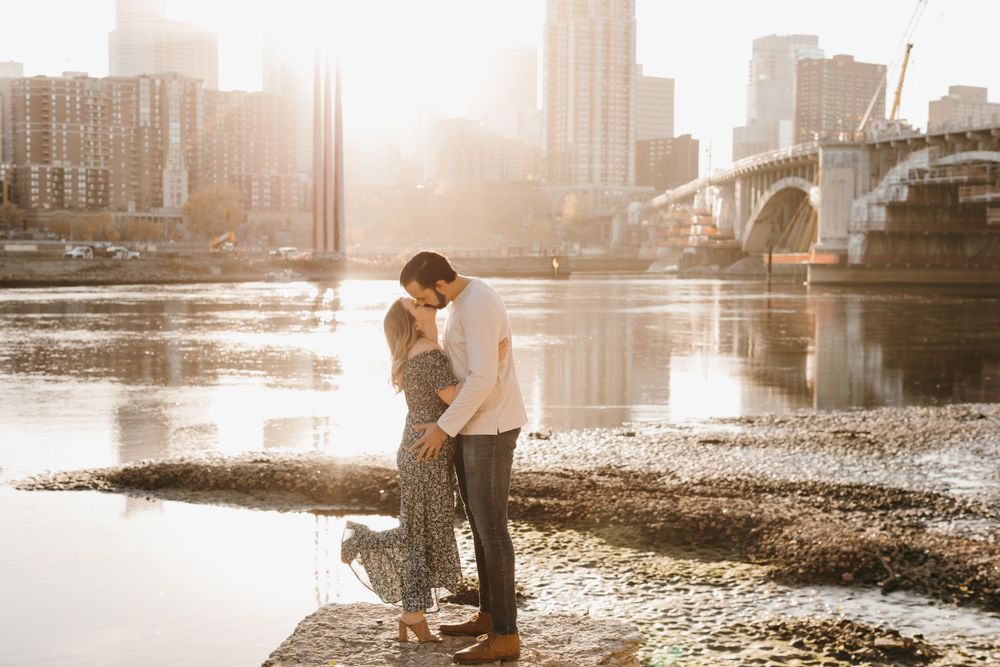 Couple sharing a tender kiss by a sunlit river with a bridge and city skyline in the background.