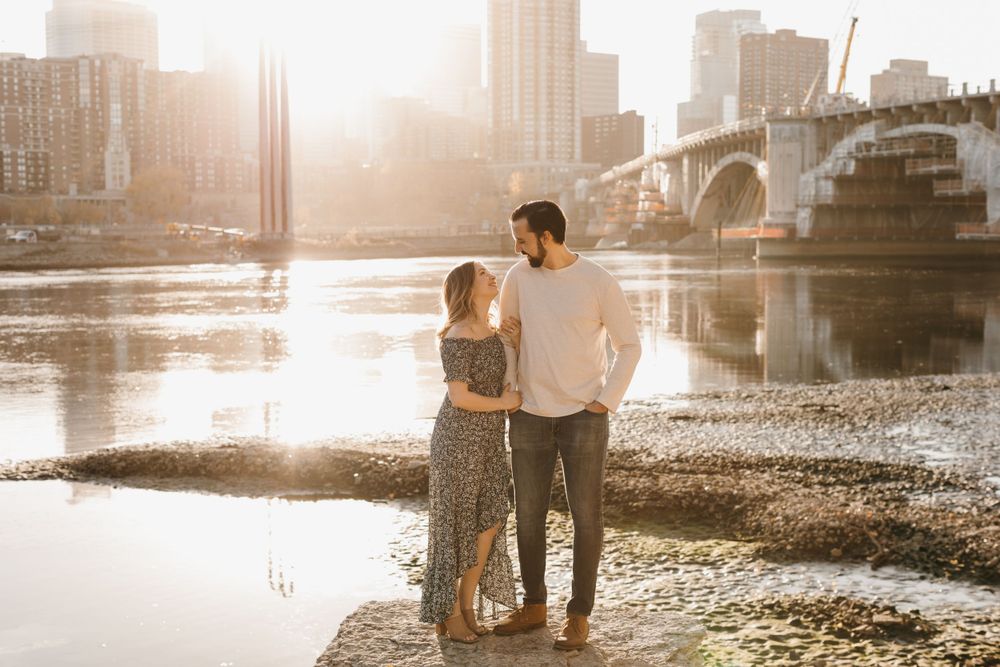 Couple holding hands and gazing at each other by a sunlit river with a city bridge in the background.