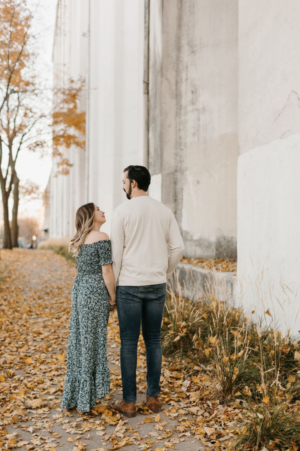 Couple holding hands while gazing at each other beside a white building in fall.