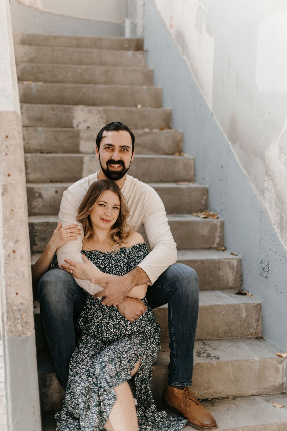 A bearded man hugs a smiling woman in a floral dress while sitting on outdoor concrete steps.