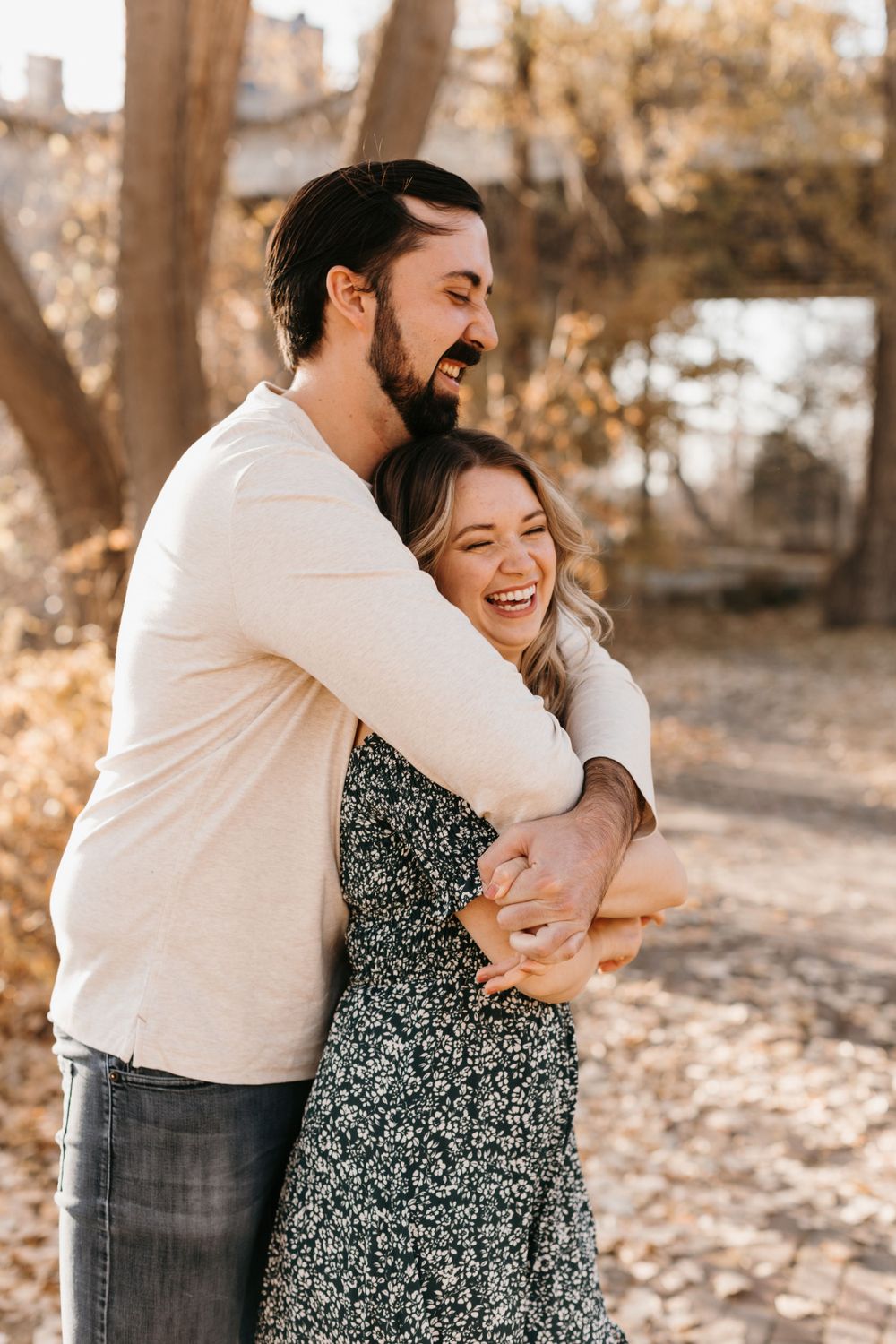 Man hugging woman from behind as she laughs joyfully outdoors in autumn, both wearing casual fall outfits.