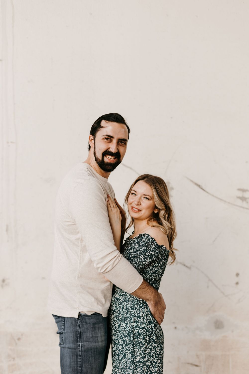 Playful couple posing against a white wall, woman leaning into man who smiles at the camera.