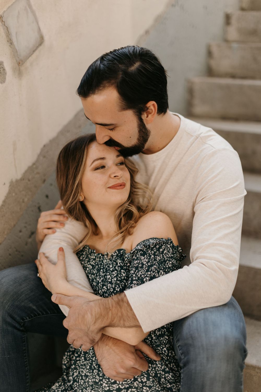 A bearded man gently kisses a womans forehead while embracing her on concrete outdoor stairs.