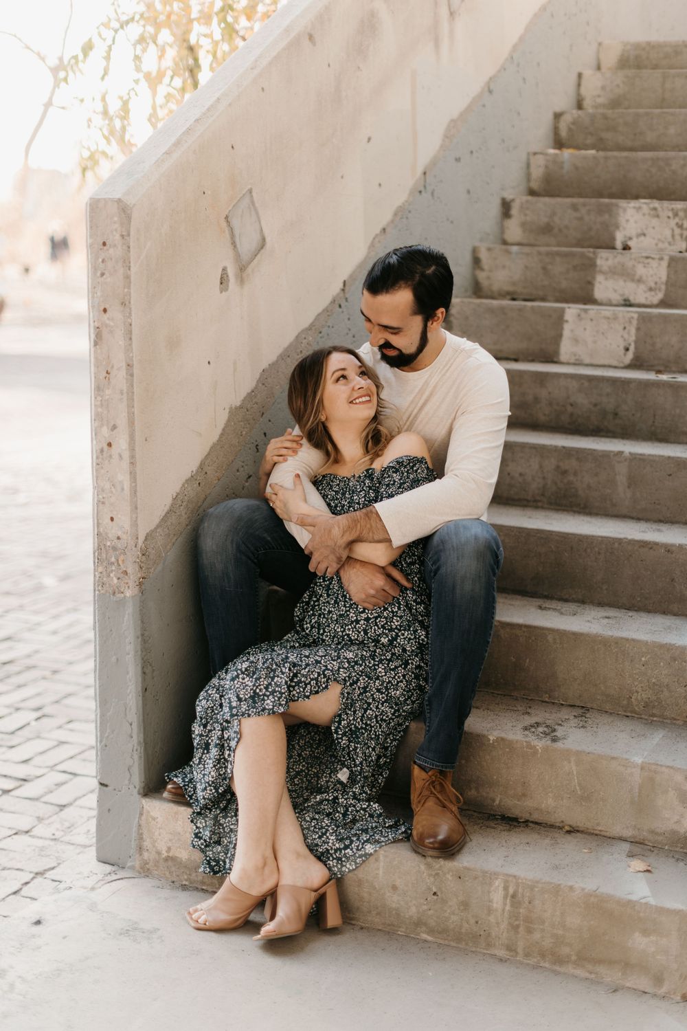 A laughing woman in a floral dress sits between a mans legs on concrete steps as he holds her.