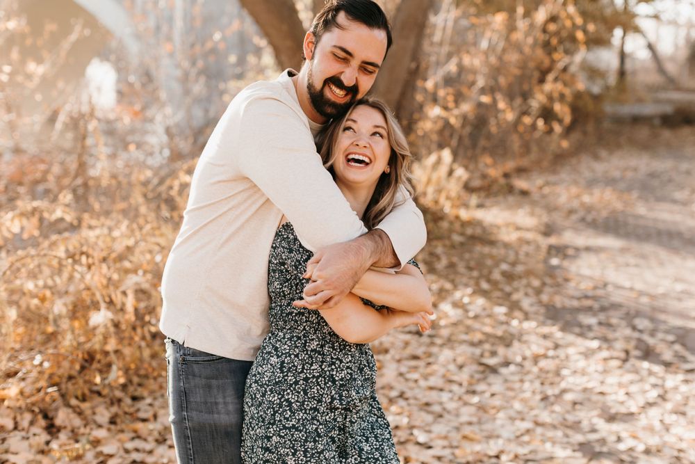 Joyful couple laughing together outdoors, man hugging woman from behind in a sunny autumn setting.