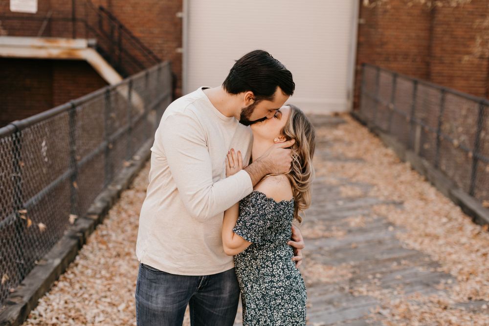 Couple sharing a tender kiss outdoors, man in white sweater embracing woman in dark floral dress in autumn setting.