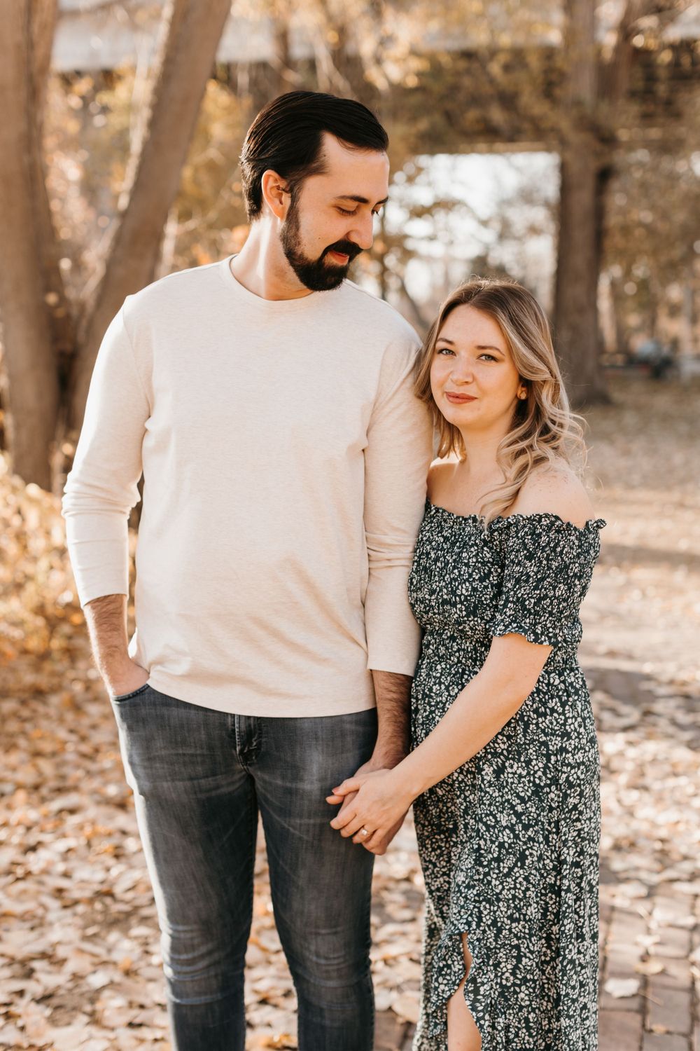 Couple holding hands outdoors in autumn park, man in beige sweater looking down at woman in dark floral dress.