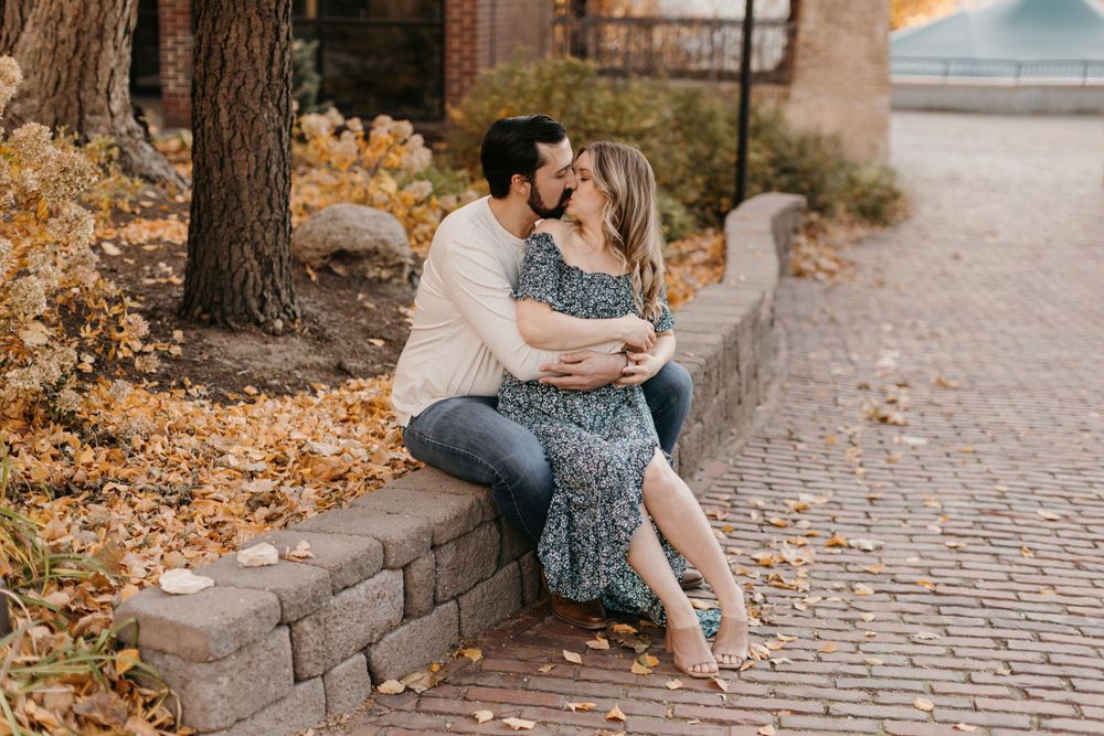 Couple sharing a kiss on a stone ledge with vibrant fall foliage backdrop.