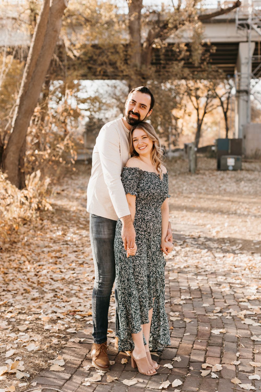 Smiling couple posing together on a brick path holding hands in a sunny fall outdoor setting.