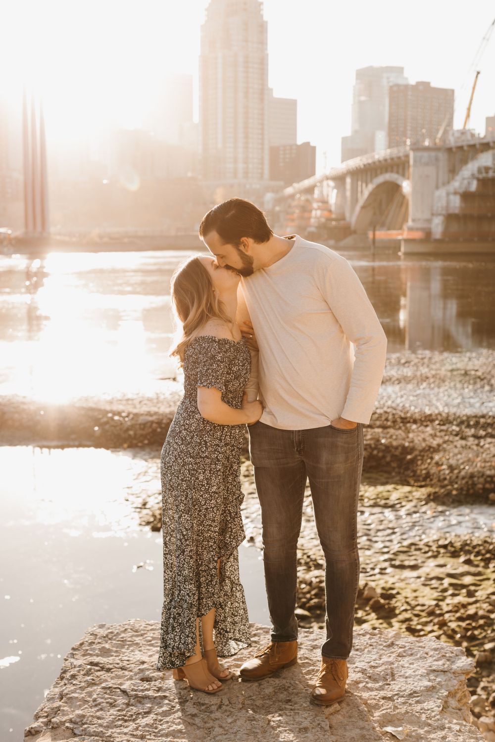 Couple sharing a kiss by a shimmering river at sunset with a city bridge and skyline behind them.