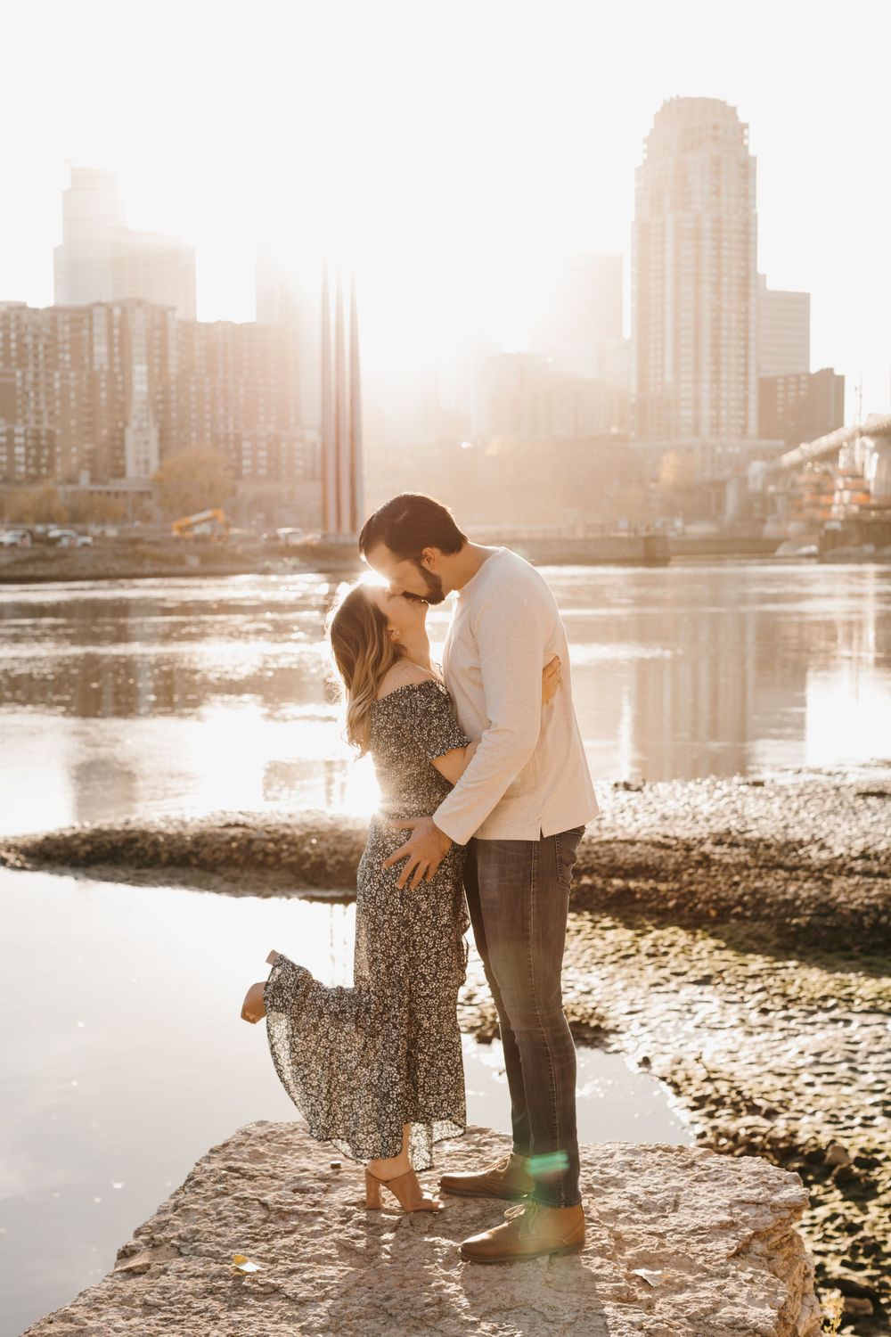 Couple kissing at golden hour by a river with city skyline, woman in floral maxi dress and man in white shirt.