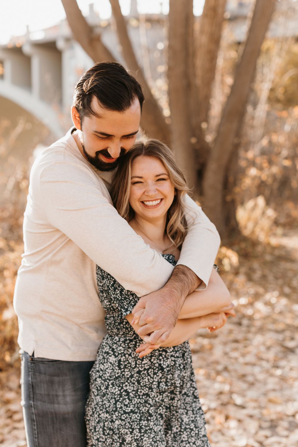 Couple embracing outdoors in autumn, woman laughing joyfully as man hugs her from behind near bare trees.