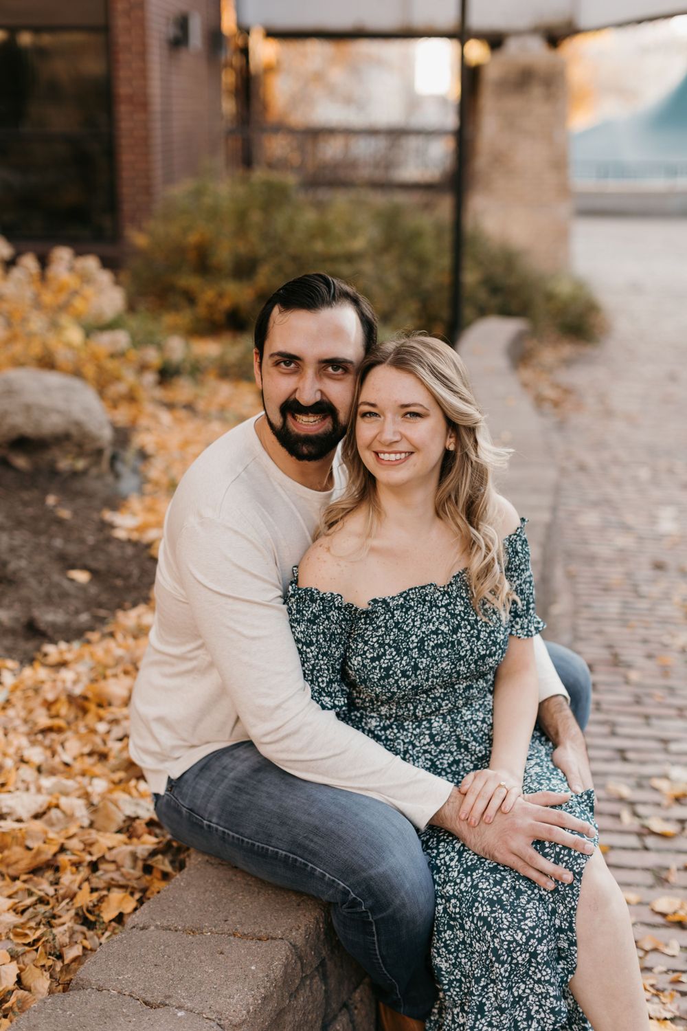 Couple smiling and embracing on a stone ledge surrounded by golden autumn leaves.