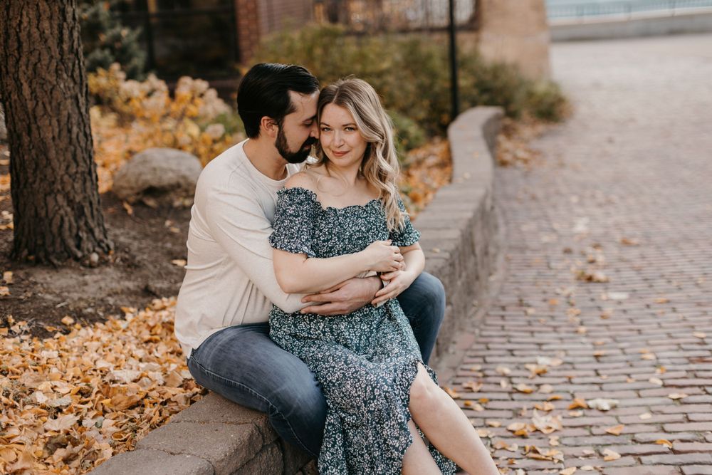Couple sitting close together on a stone ledge surrounded by golden autumn leaves.