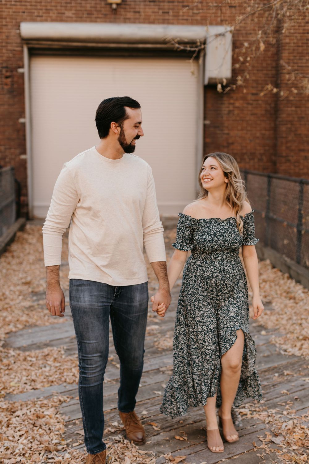 Couple holding hands and walking together outdoors in fall, man in cream sweater and woman in floral dress smiling.