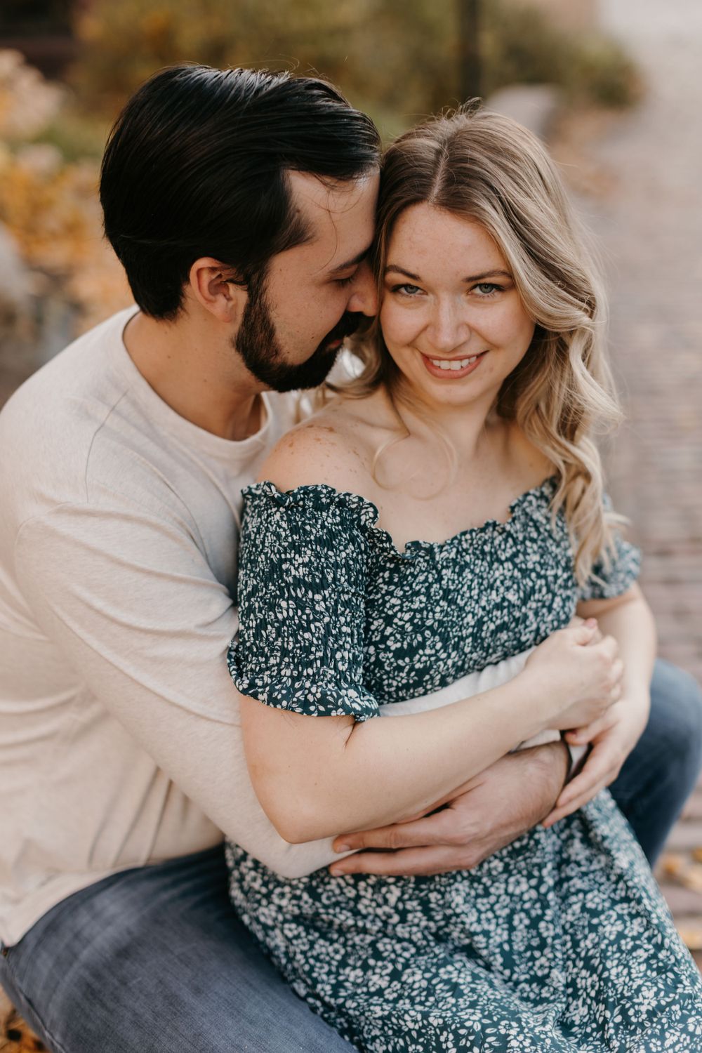 Man embracing woman in floral dress as she smiles during fall engagement photos.