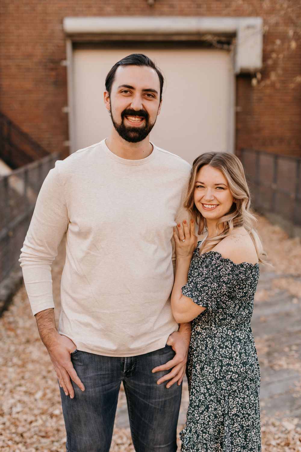 Happy couple posing together outdoors, man in beige sweater and woman in dark floral off-shoulder dress smiling at camera.