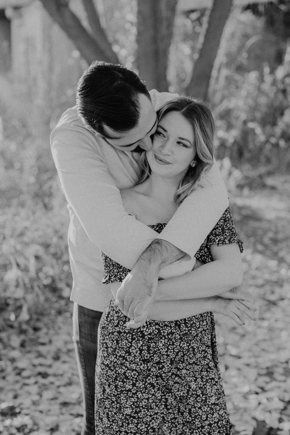 Black and white photo of couple embracing tenderly, man nuzzling woman's cheek among autumn trees.