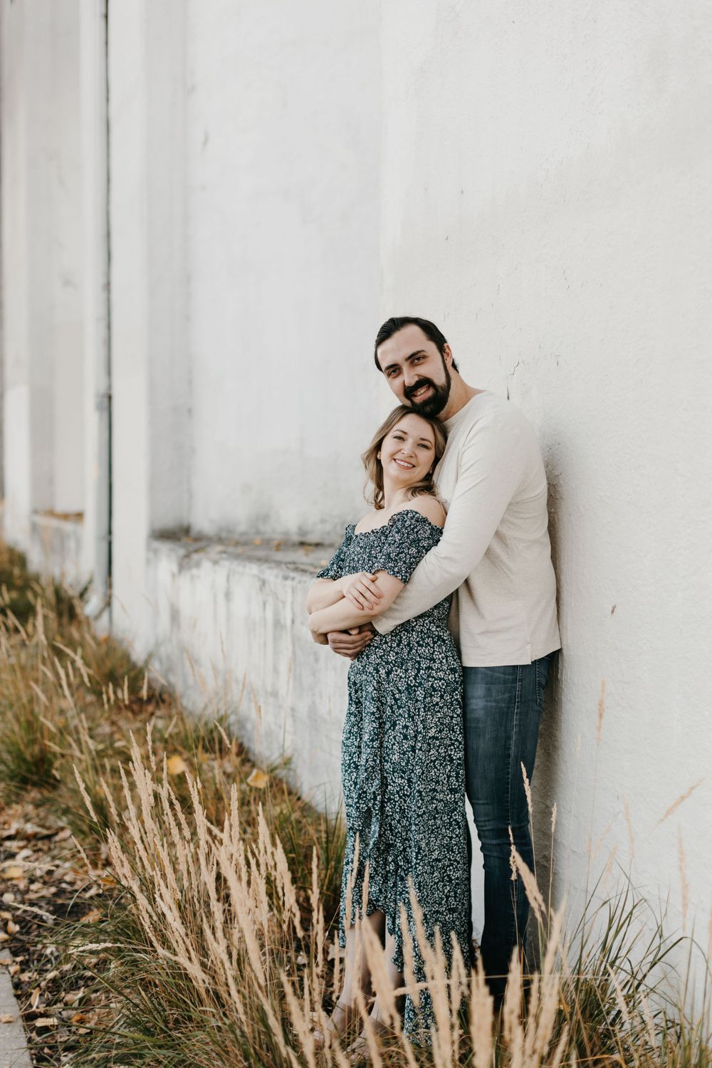 Couple smiling and embracing against a white wall surrounded by tall ornamental grasses in autumn.