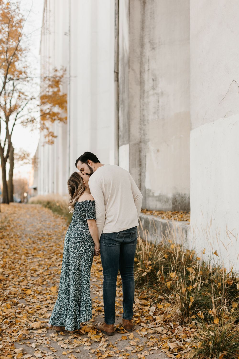 Couple sharing a romantic kiss while holding hands along a leaf-covered autumn path.