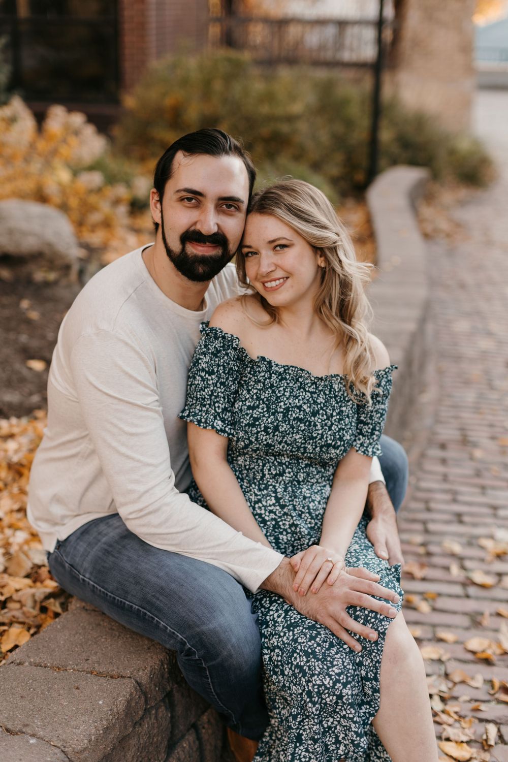 Happy couple cuddling on a stone wall with colorful autumn leaves in background.