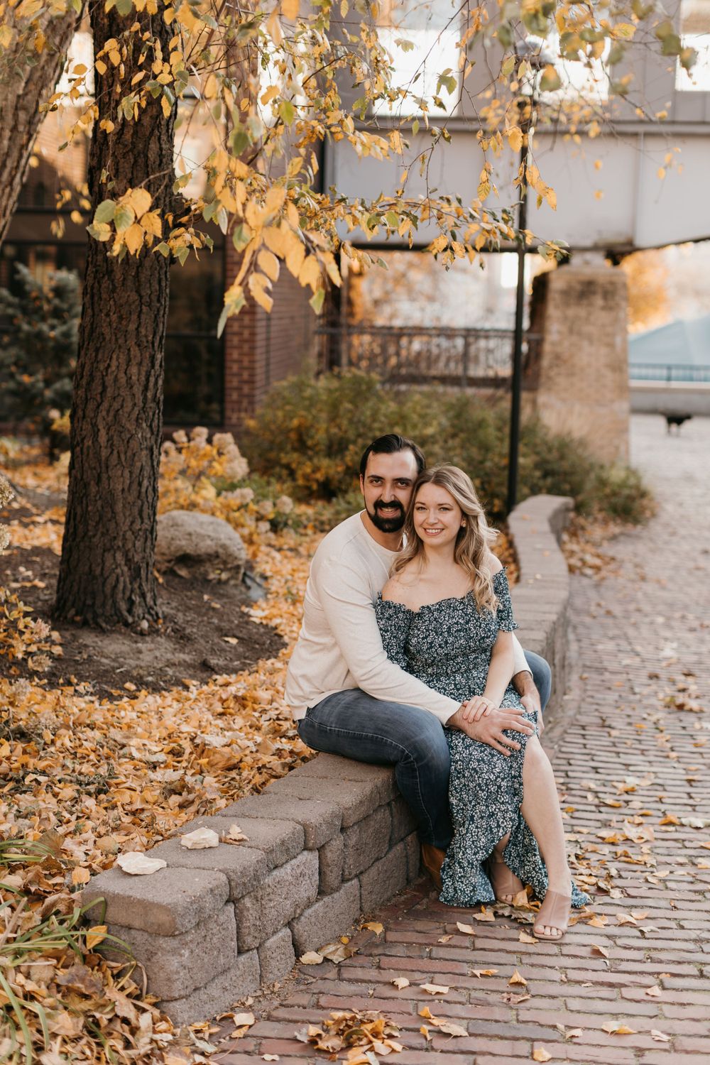 Couple sitting on a stone ledge under autumn trees with golden fallen leaves and a brick pathway nearby.