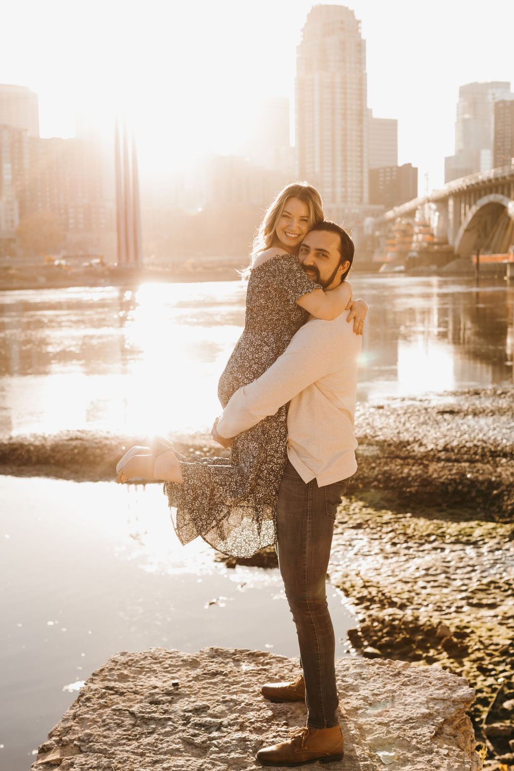 Man lifting woman in a floral dress by a golden-lit river with a city skyline and bridge in the background.