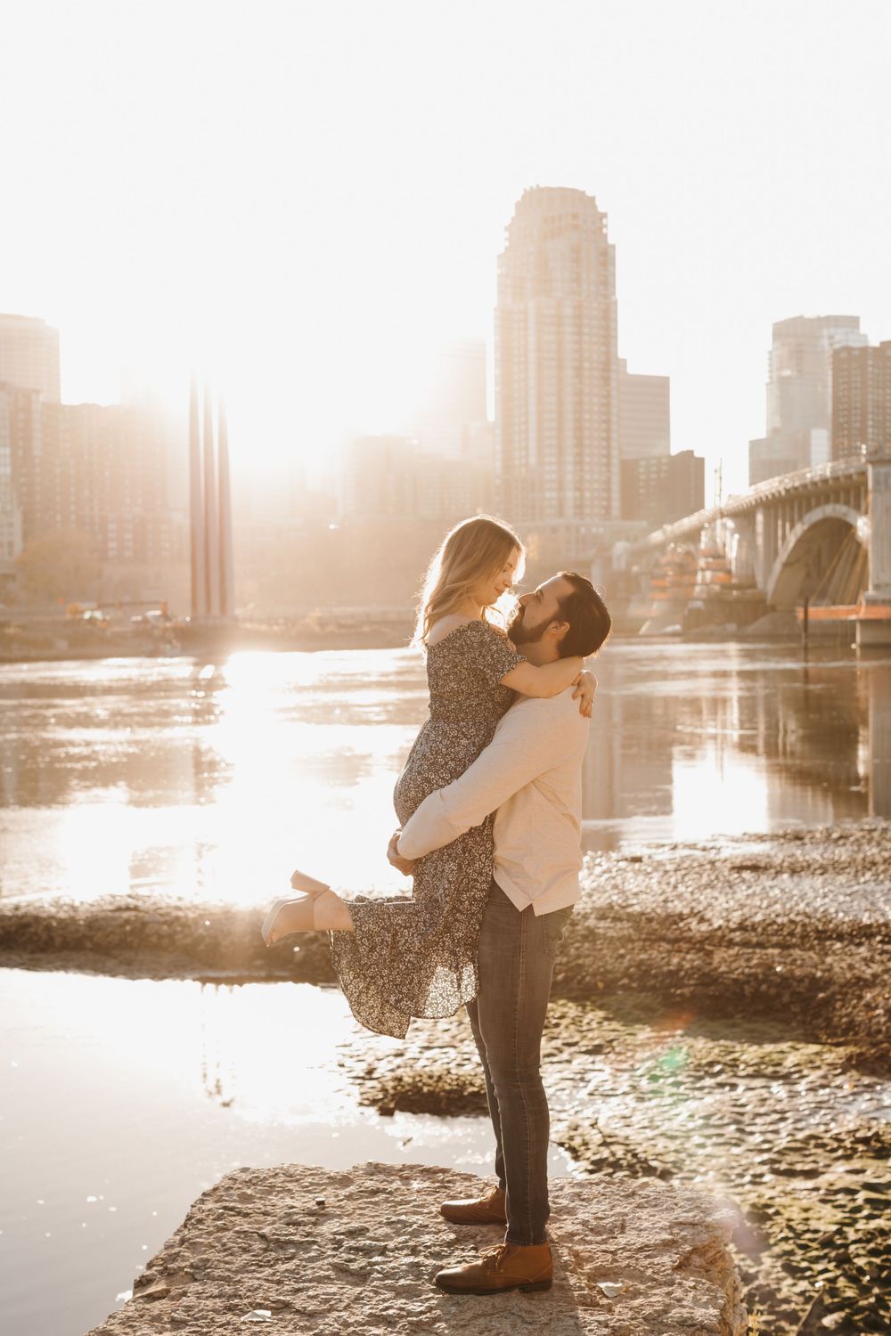 Man lifting woman for a kiss at golden hour by a river with a city skyline and bridge behind them.
