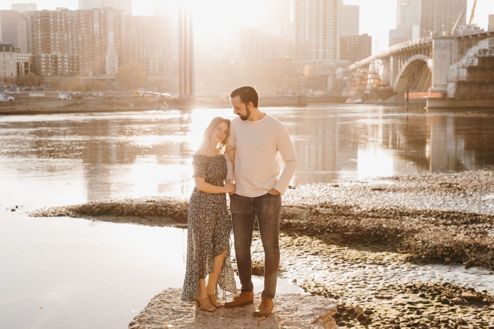 Couple standing close together by a glowing river, woman in floral dress and man in white shirt smiling at each other.