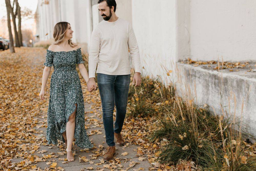 Couple holding hands walking on a leaf-covered path in autumn, woman in floral dress, man in jeans and cream sweater.