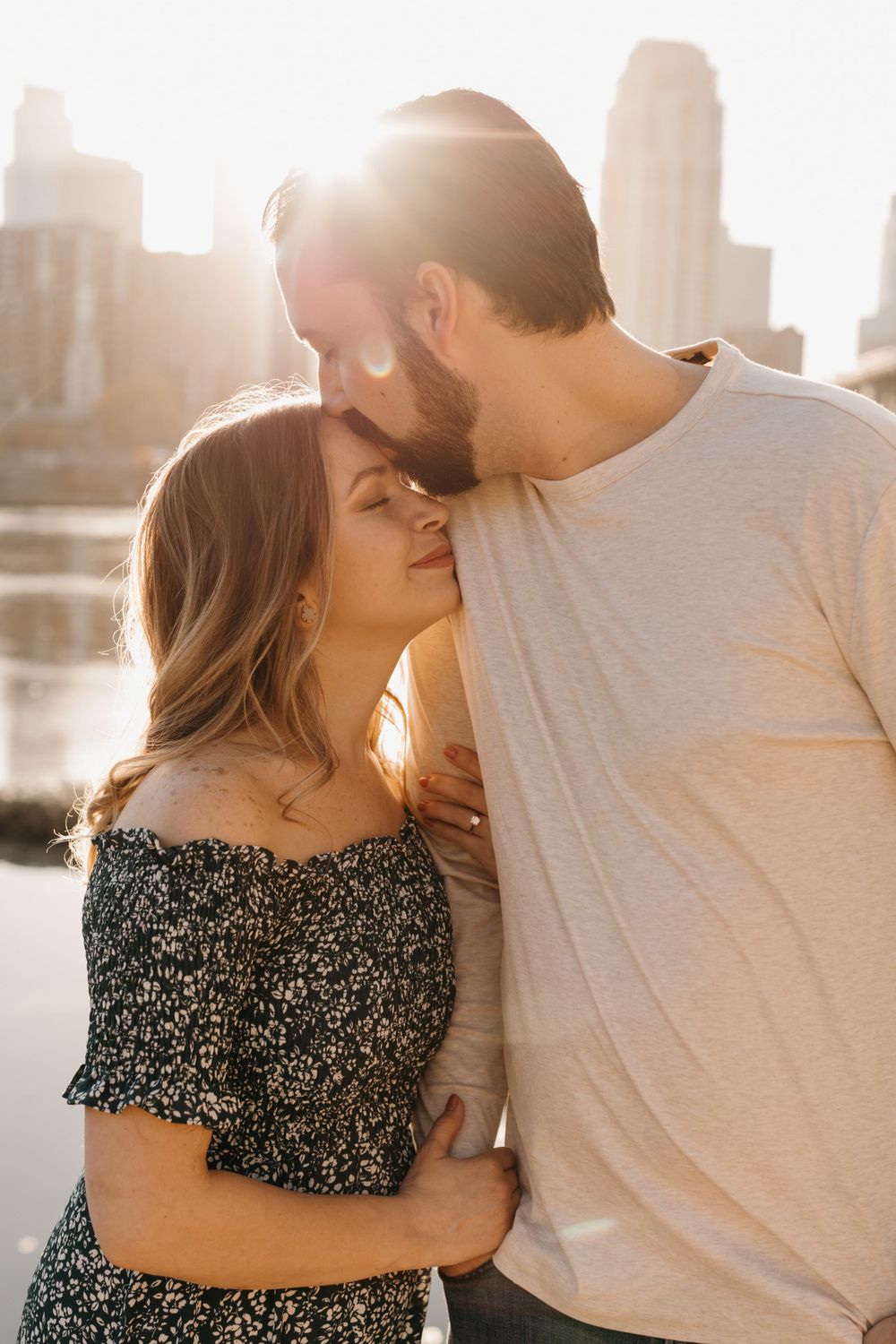 Man kissing woman on the forehead at golden hour by a river, woman in floral off-shoulder dress smiling softly.