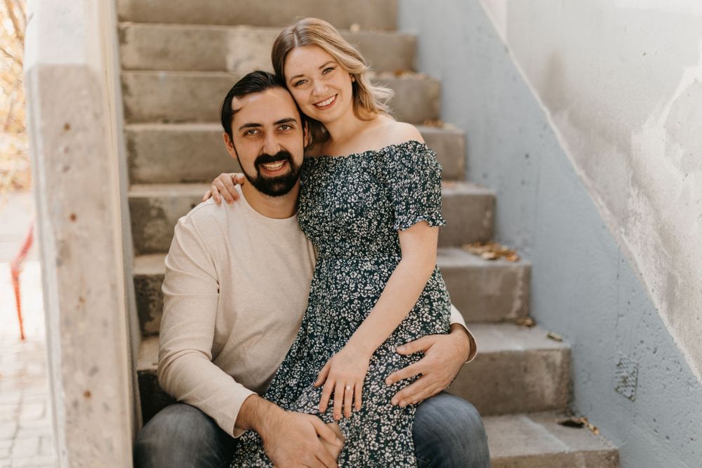 A happy couple sits close together on outdoor concrete stairs, both smiling at the camera.