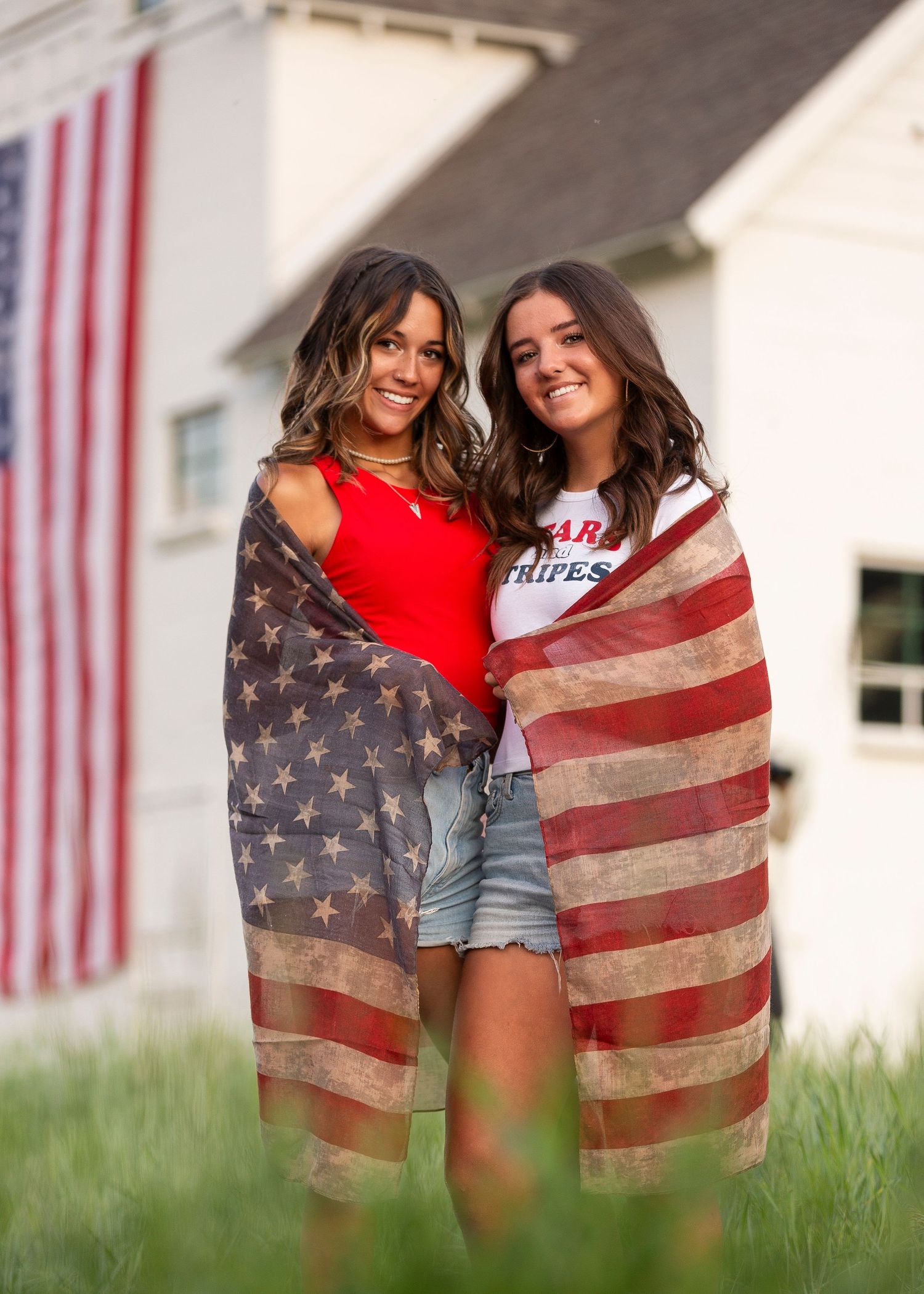 Two high school senior girls posed in front of white barn in park city Utah wrapped in American flag scarf smiling