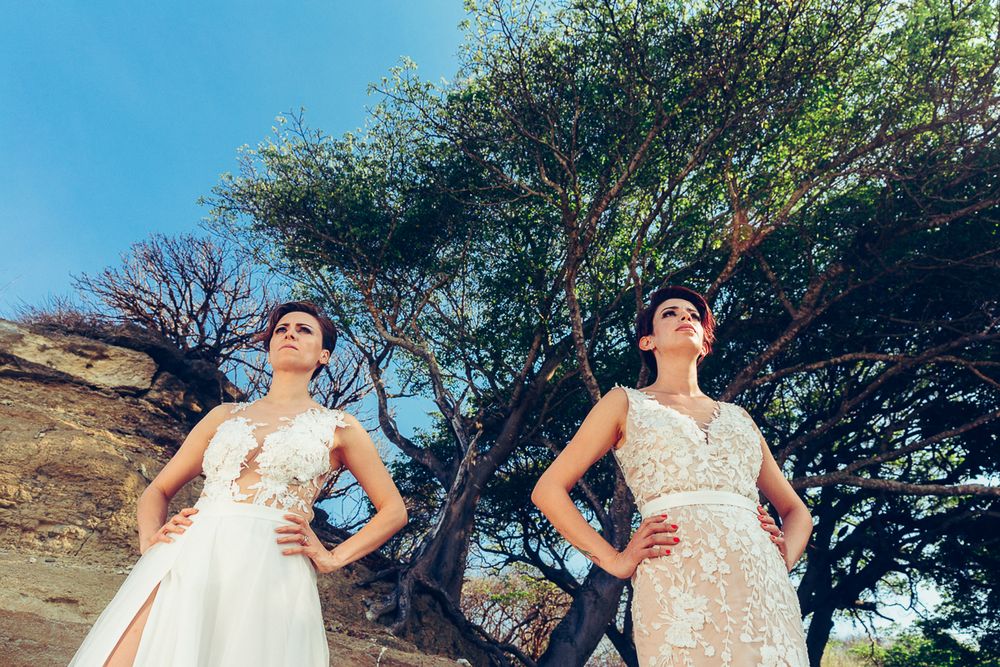 Two people in wedding dresses posing dramatically against trees and blue sky.