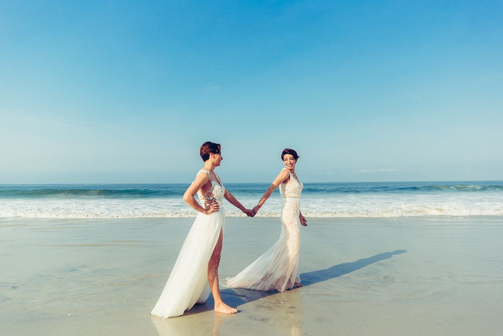Two people in elegant white dresses holding hands on a sunlit beach.