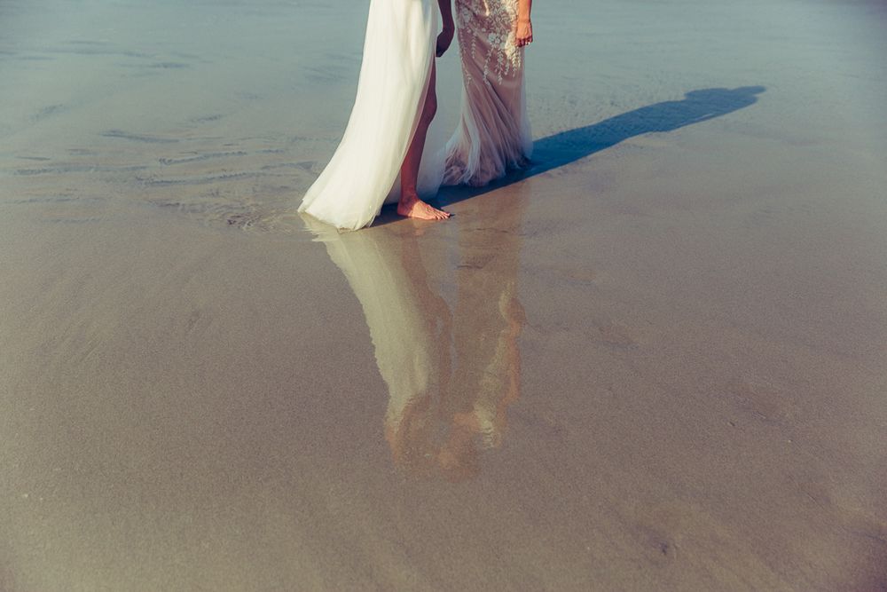 Reflections in wet sand of flowing dresses during beach photoshoot.