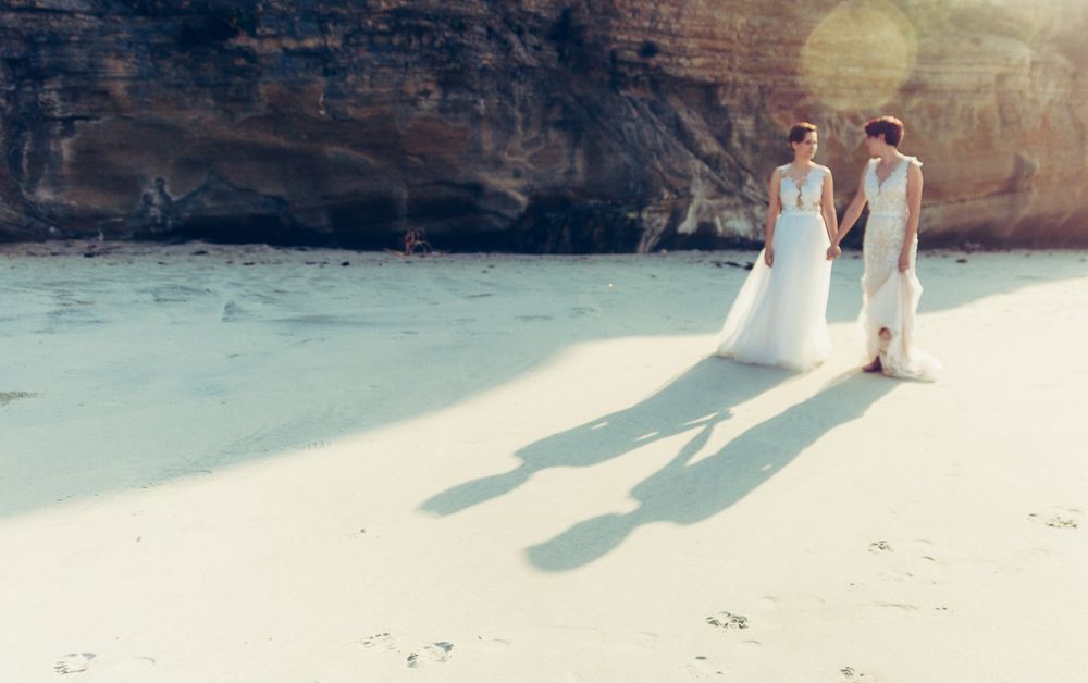 Couple in white dresses casting long shadows while walking on a pristine beach near rocky cliffs.