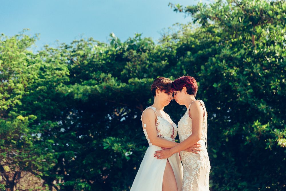 Two people in wedding dresses embracing in front of green foliage.