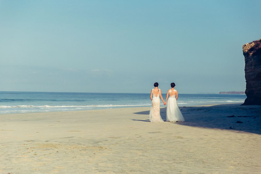 Two people in white dresses walking along a scenic beach with cliffs and blue ocean in the background.