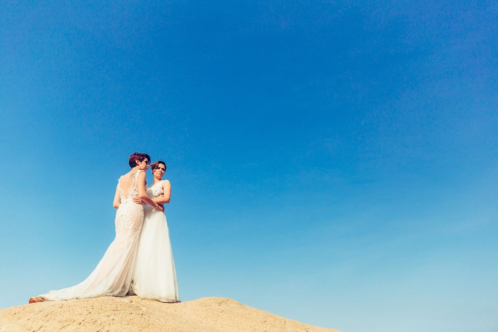 Couple in white dresses standing against bright blue sky on sandy hill.
