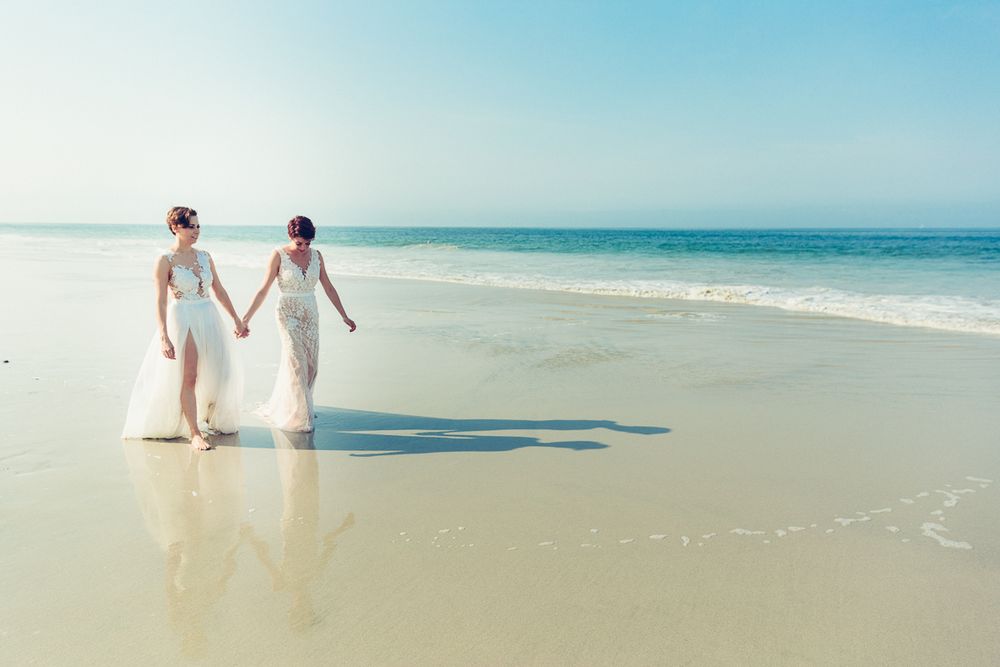 Two people in white dresses walk hand in hand along a pristine beach shoreline.