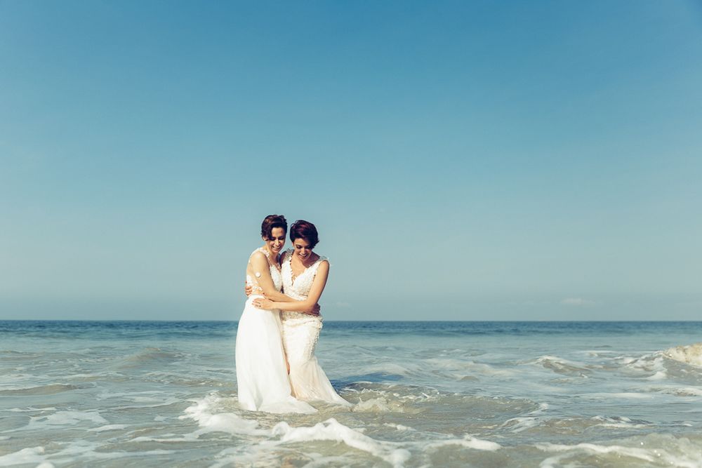 Two brides in white dresses embrace while standing in ocean waves at a beach wedding.