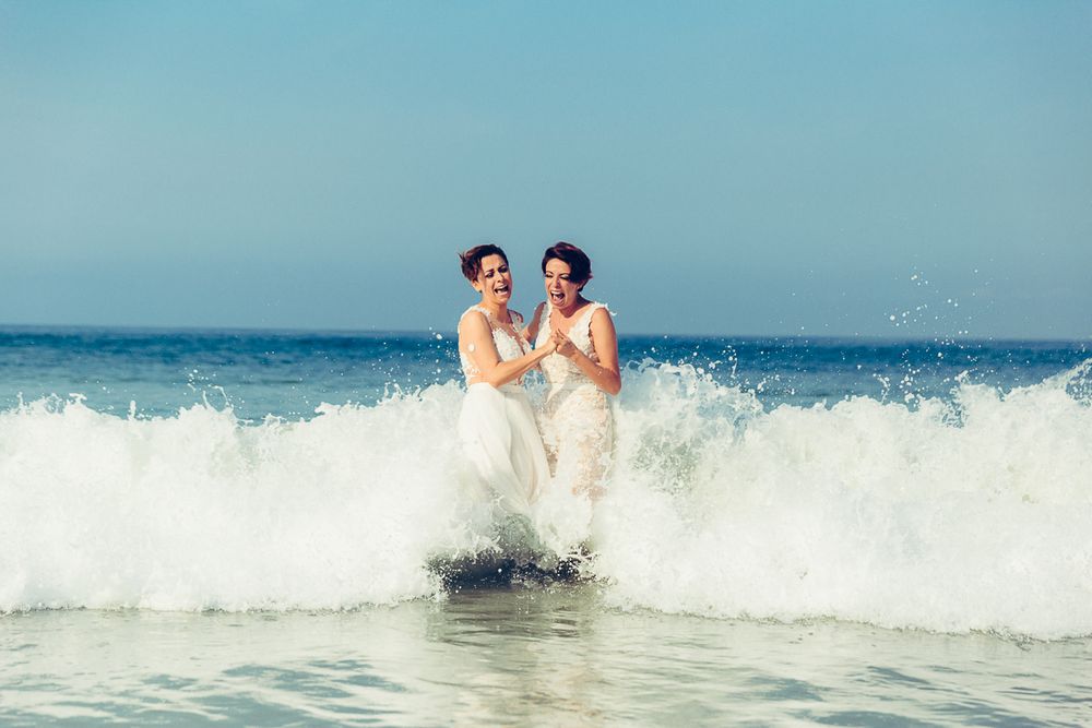Two people in white dresses laugh together as ocean waves crash behind them on a beach.