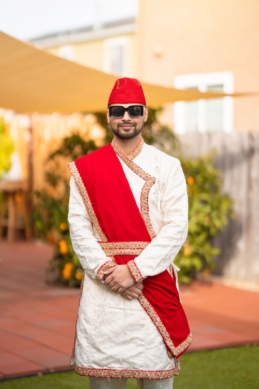 A person wearing traditional South Asian attire with a red sash and cap poses in a garden setting.