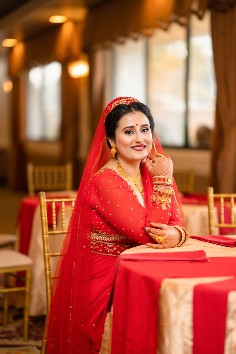 A bride in a red embroidered sari leans gracefully against a reception table with golden chairs.