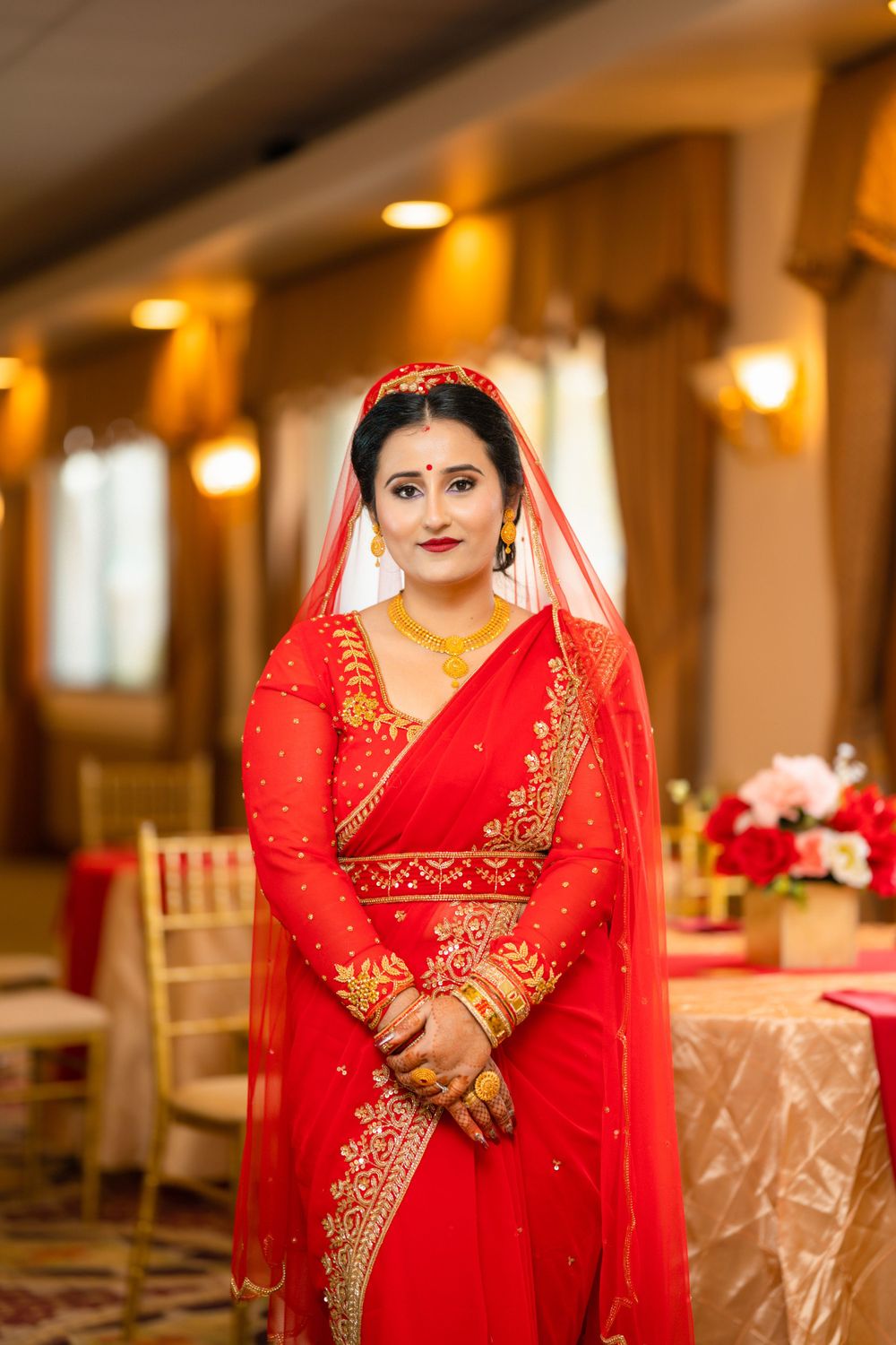 A bride poses in a stunning red and gold sari with traditional jewelry at an indoor wedding venue.