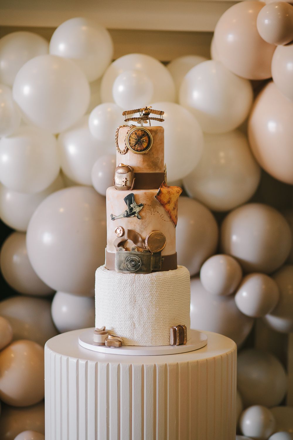 a decorated baby shower cake white and brown with traveling items like a camera and a plane with a balloon background
