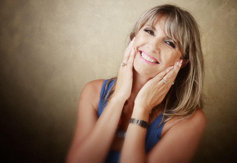 Joyful portrait of woman in blue top with hands on cheeks against warm background.